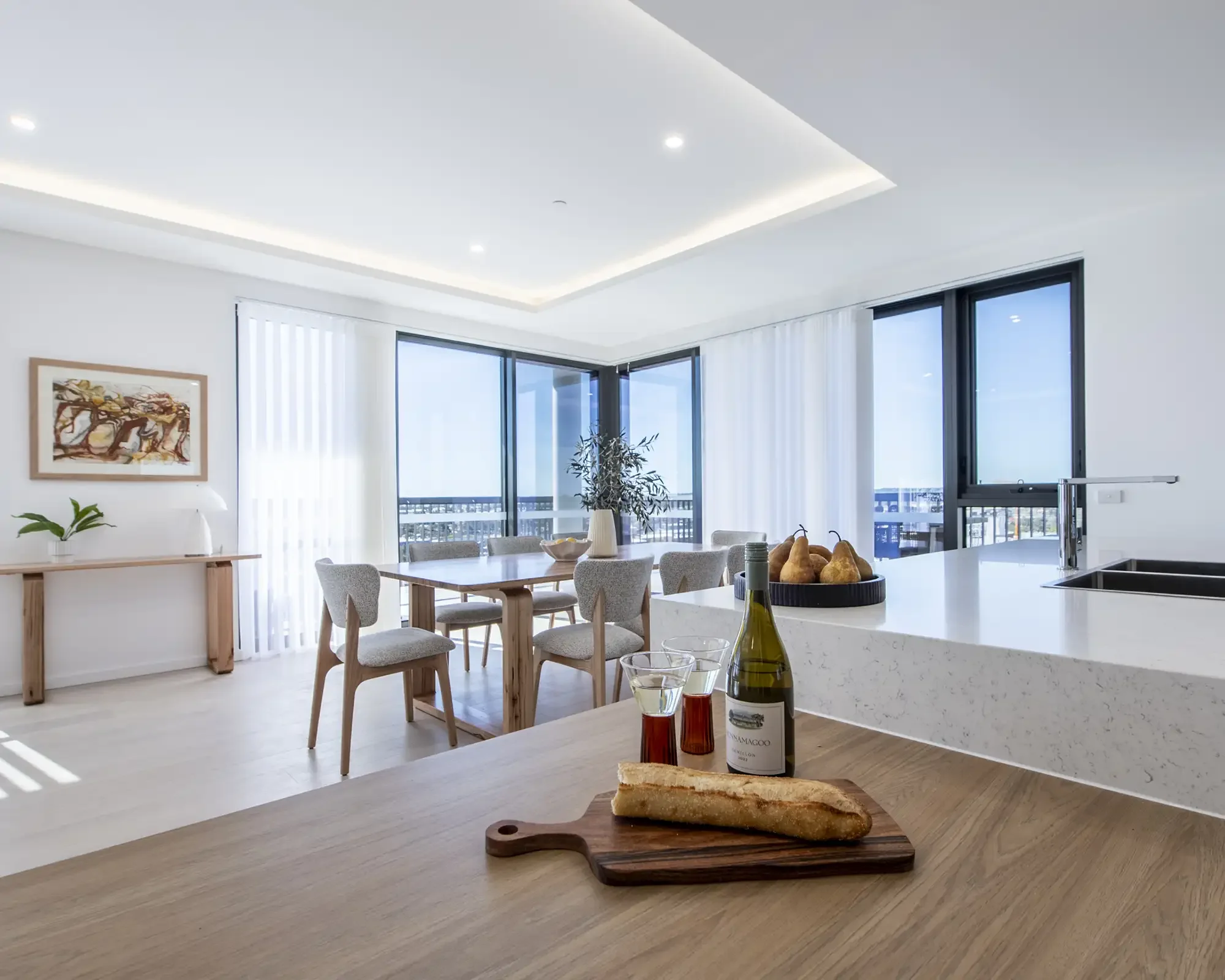 Modern kitchen with a white island, wooden table, and chairs. Large windows with white curtains. Ceiling with recessed lighting. Artwork on the wall, plants, and food items like bread and wine on the counter and table.