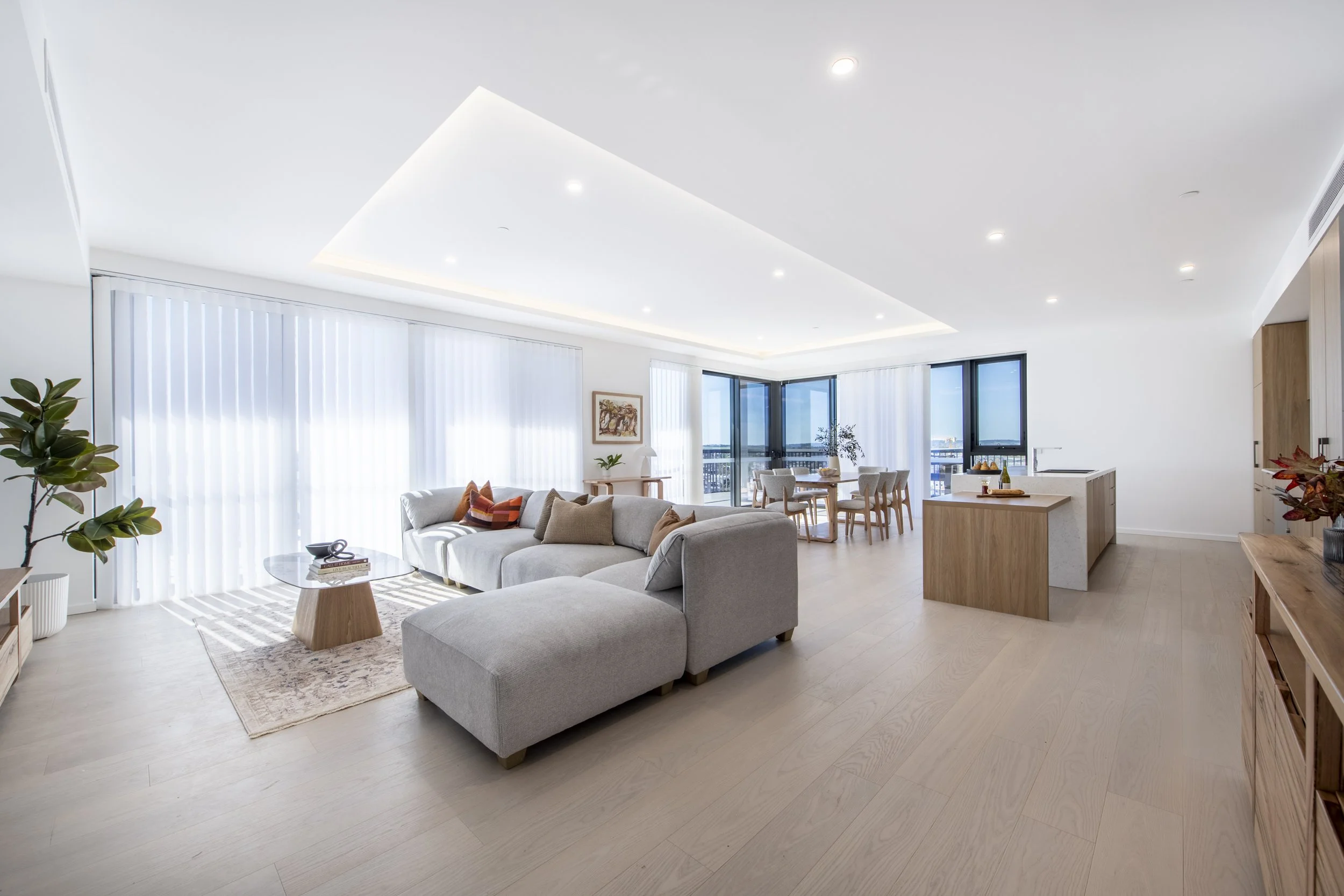 Bright, modern open-plan living room and dining area with white walls, large windows, light wood flooring, gray sofa, round coffee table, plants, and a kitchen island with chairs.