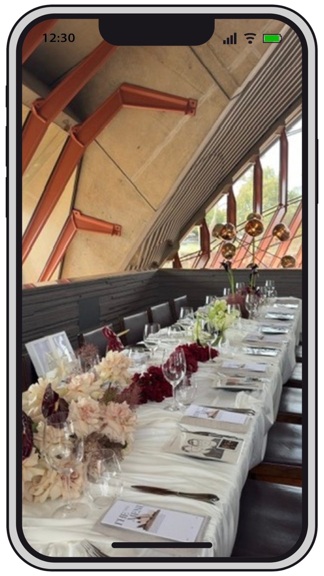 A long dining table set for a formal meal with a white tablecloth, floral centerpieces, wine glasses, utensils, and printed menus, arranged in a room with sloped ceilings, large windows, and decorative lighting.