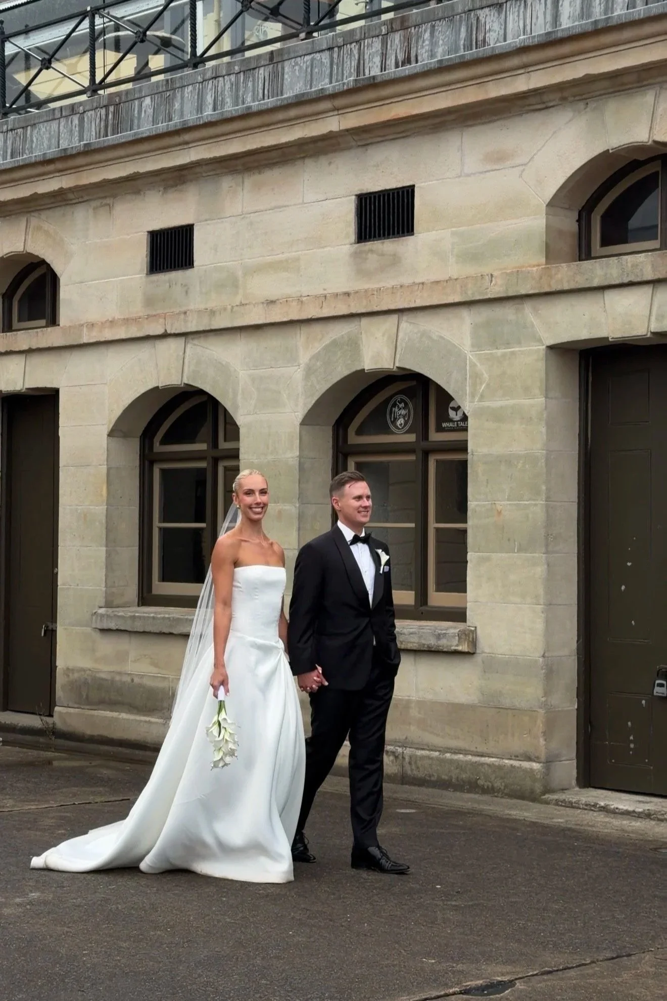 A bride and groom holding hands, standing outside in front of a stone building with arched windows, dressed in wedding attire, smiling and looking to the side.