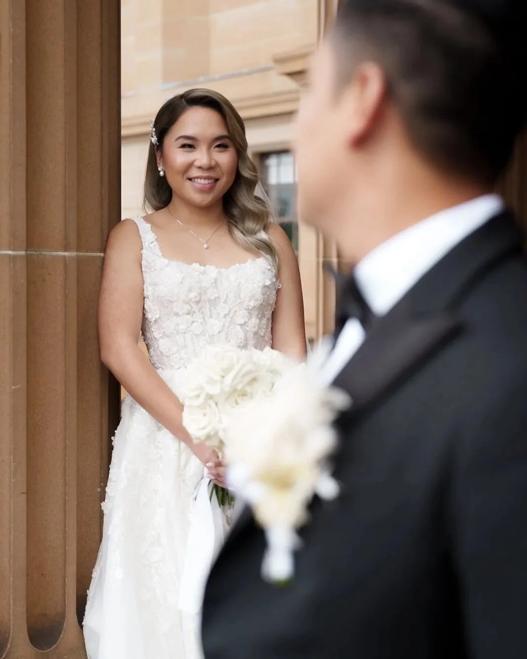 A bride in a wedding dress holding a bouquet, smiling at a groom dressed in a tuxedo, standing in front of a building with wood paneling.
