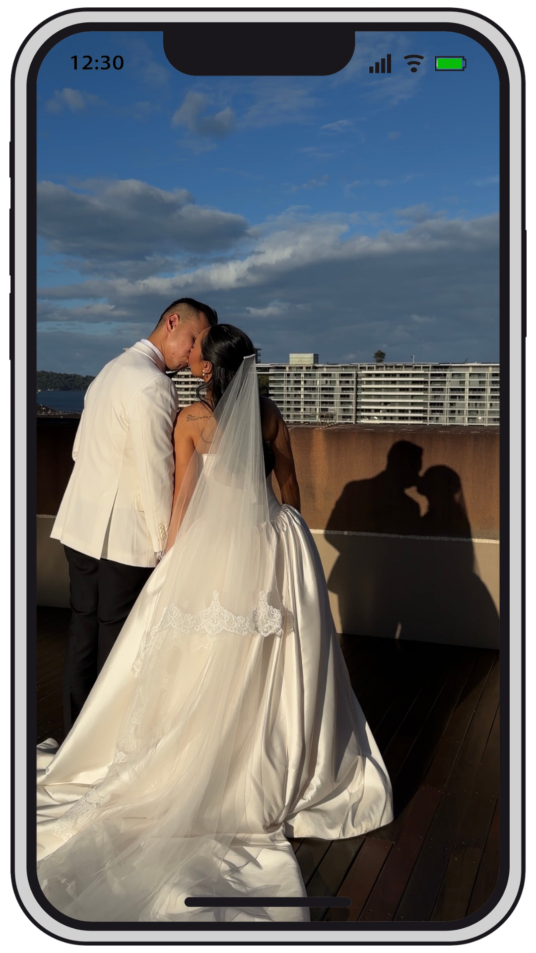 A wedding couple sharing a kiss on a rooftop balcony with a cityscape and sky in the background, casting romantic shadows on the wall.