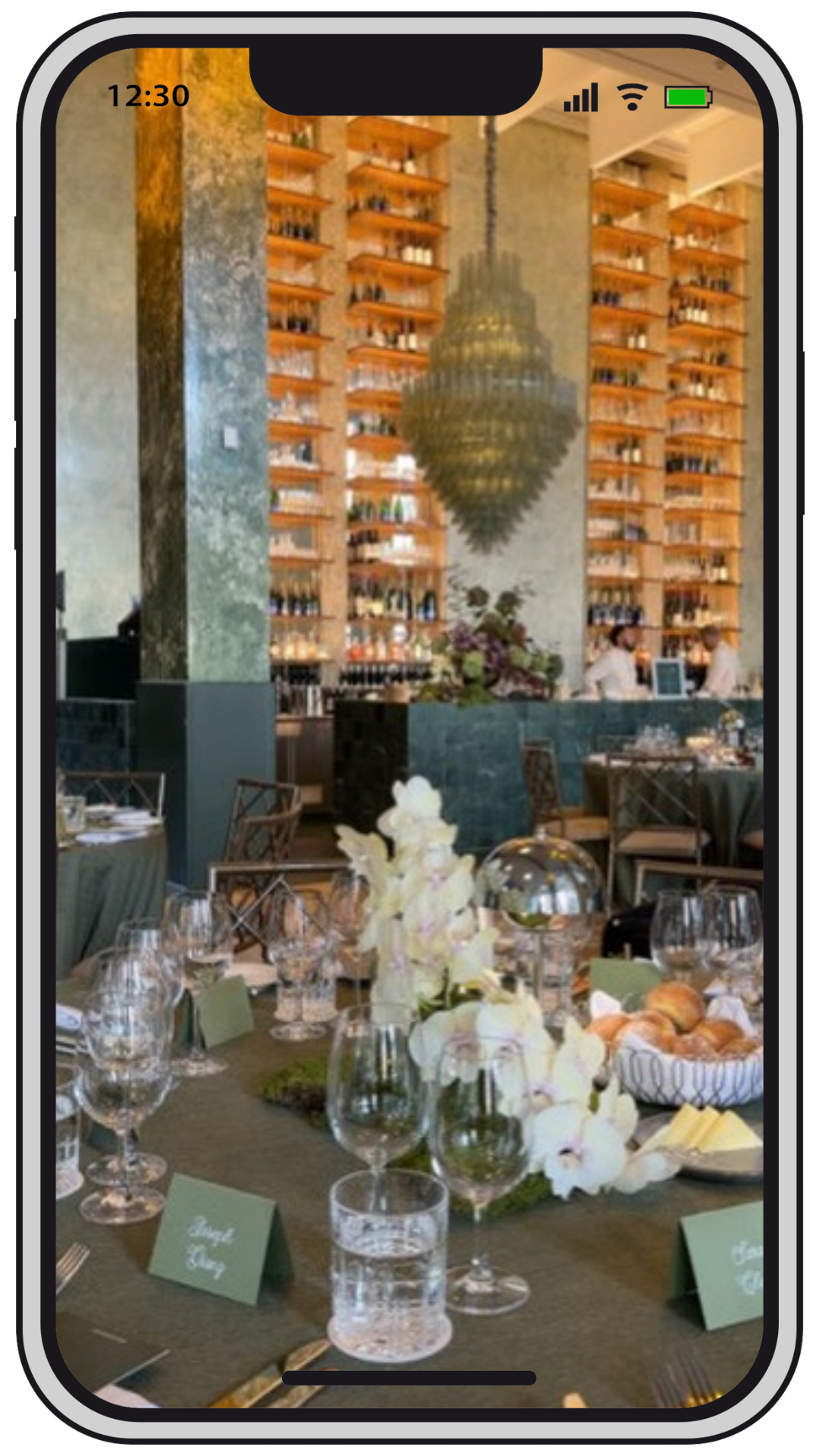 Elegant restaurant interior with a floral centerpiece, glassware, and place cards on a dining table, with a large decorative chandelier and shelves filled with bottles in the background.