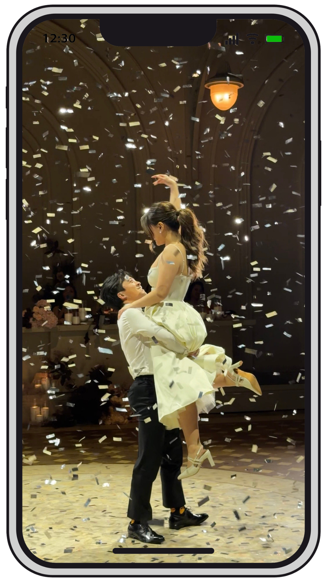 A man and woman dancing amidst falling confetti in a decorated indoor venue, with the woman lifted in the air and both smiling.