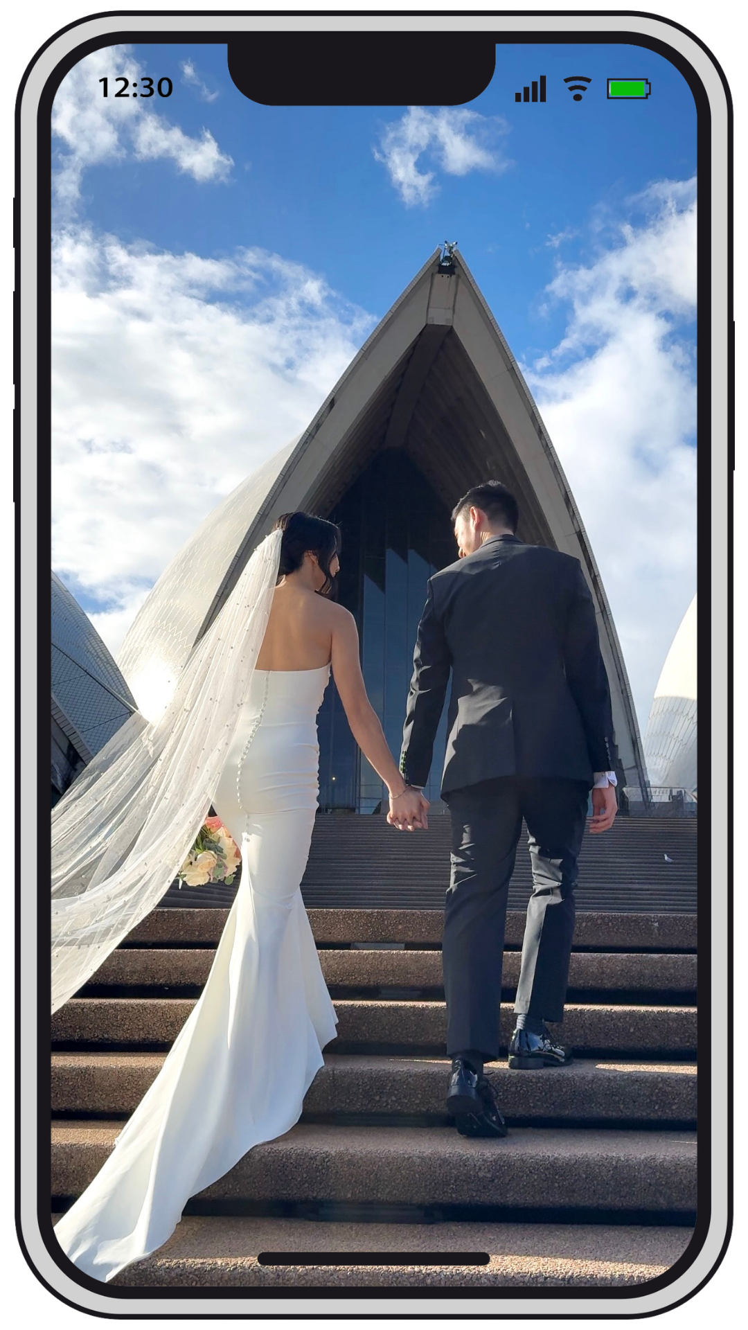 A bride and groom holding hands while walking up steps outside a modern building with an arch design, under a partly cloudy sky.