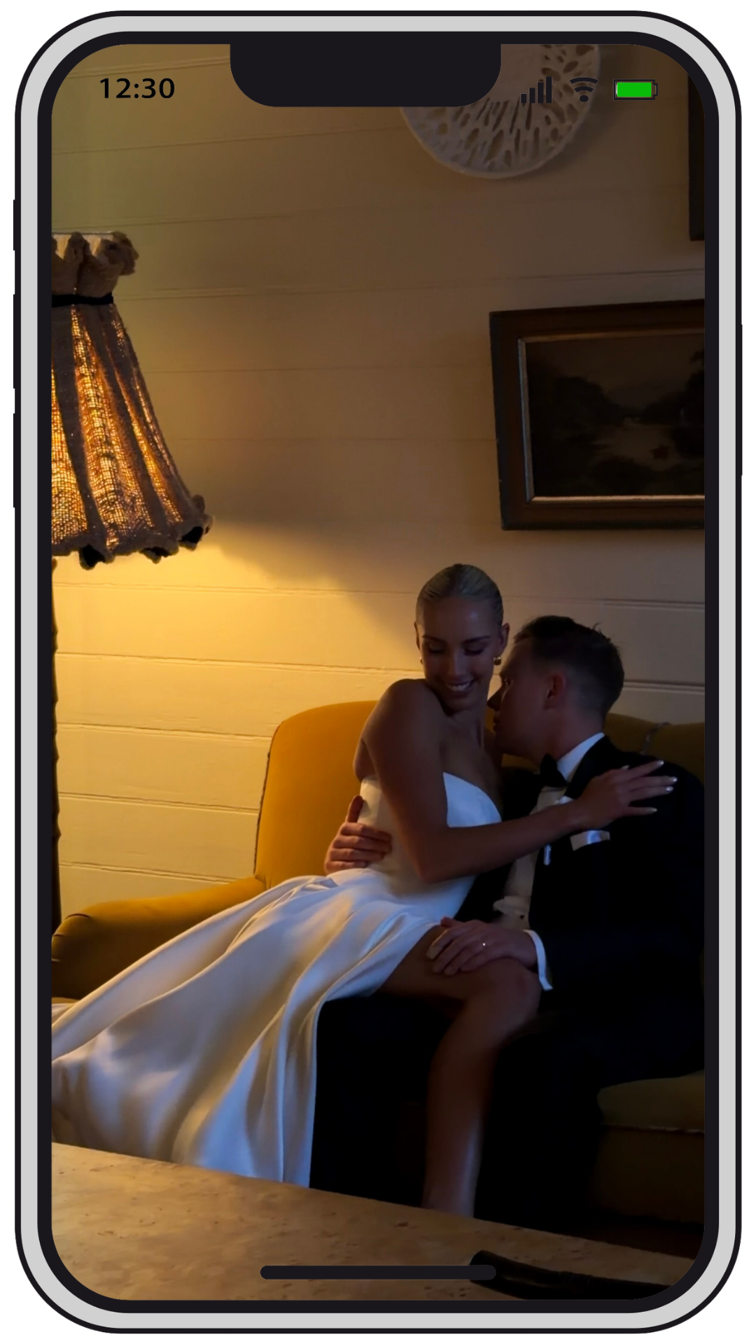A woman in a wedding dress sitting on a yellow couch with a man in a tuxedo, sharing an intimate moment in a warmly lit room with wall art and a vintage lamp.