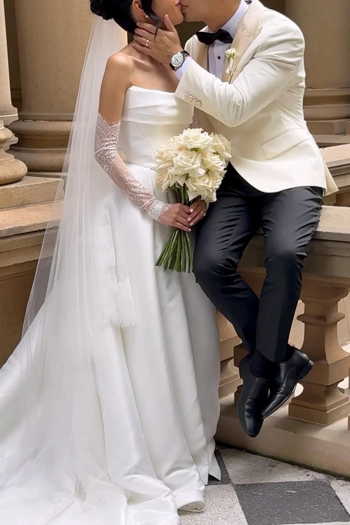 A bride and groom sharing a kiss during their wedding, with the bride holding a bouquet of white roses, where the groom is wearing a white tuxedo jacket with a black bow tie, and the bride is in a white wedding gown with lace sleeves, standing on steps outside of a stone building.