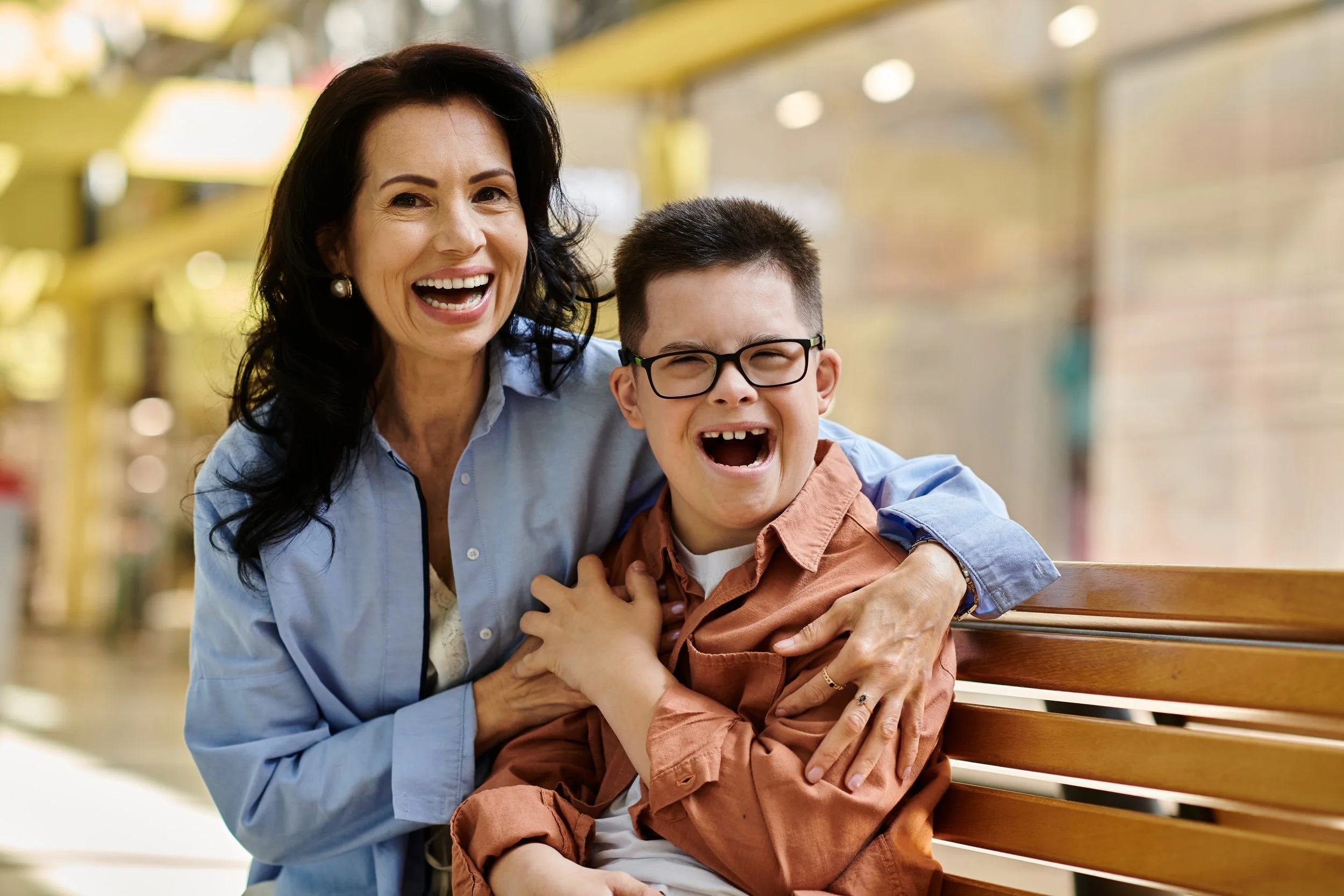 A woman and a boy sitting on a wooden bench in a brightly lit indoor space, smiling and laughing together.