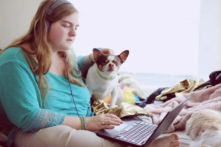 A young woman with long red hair wearing headphones sitting on a bed, using a laptop, with a small dog on her shoulder and sleeping part of a dog in the foreground.