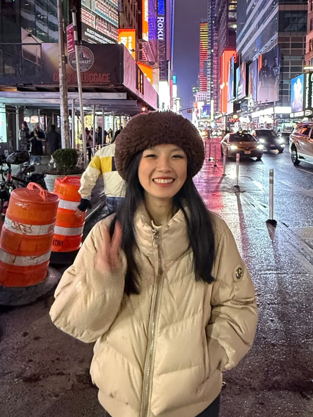 A smiling woman in a beige winter jacket and a large brown hat standing on a busy city street at night illuminated by bright neon signs, with traffic and construction barrels visible.