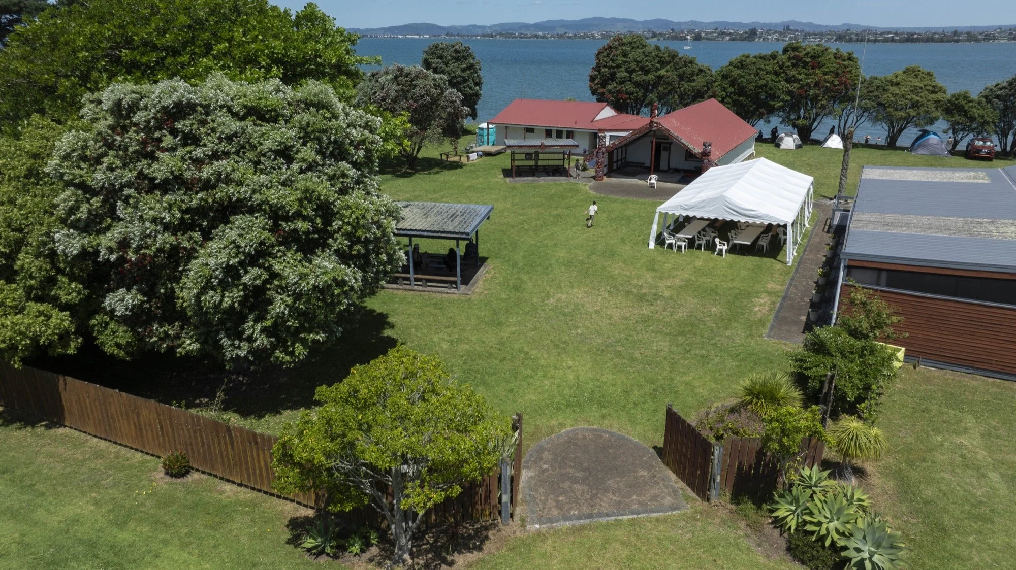 Aerial view of a lakeside lodge with red-roofed buildings, tents, event canopy, landscaped lawn, and distant hills