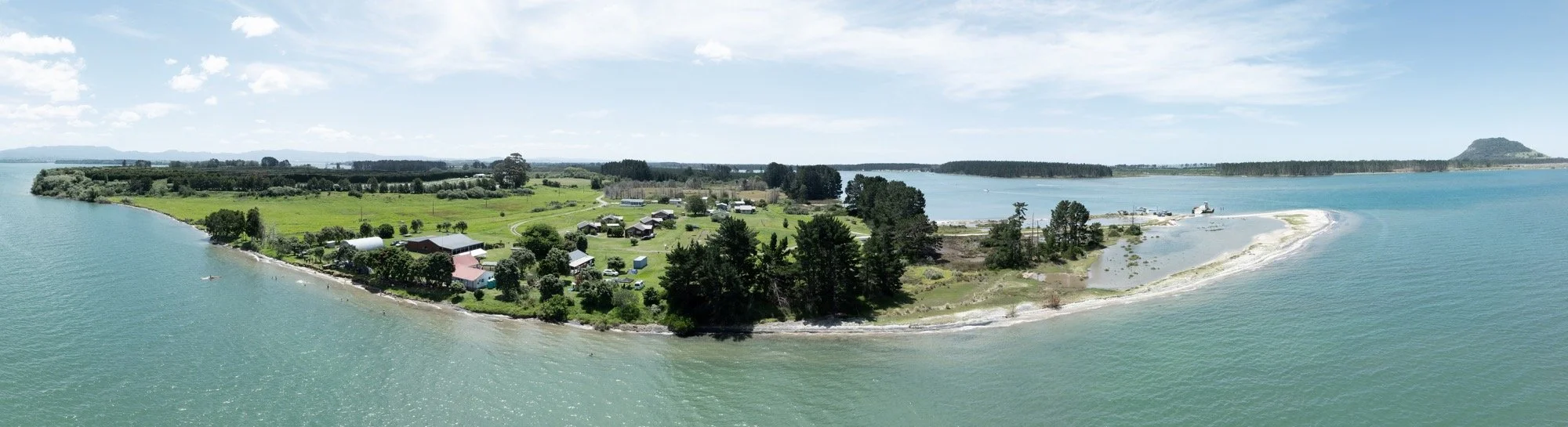 Aerial panorama of a green coastal peninsula with homes, farmland, sandy beaches, turquoise water, and a forested island under a blue sky