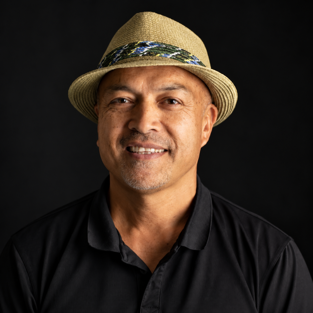 Smiling man wearing a decorative fedora and black polo shirt against a black background