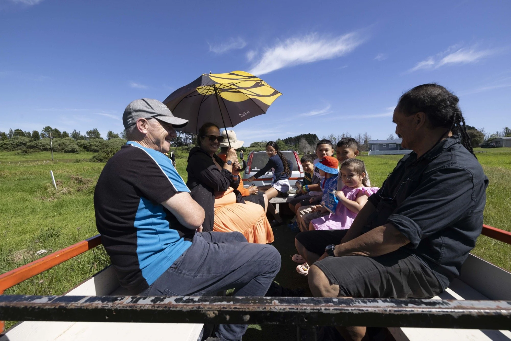 Multigenerational family riding in an open wagon through a sunny rural field, with a yellow-and-black umbrella overhead