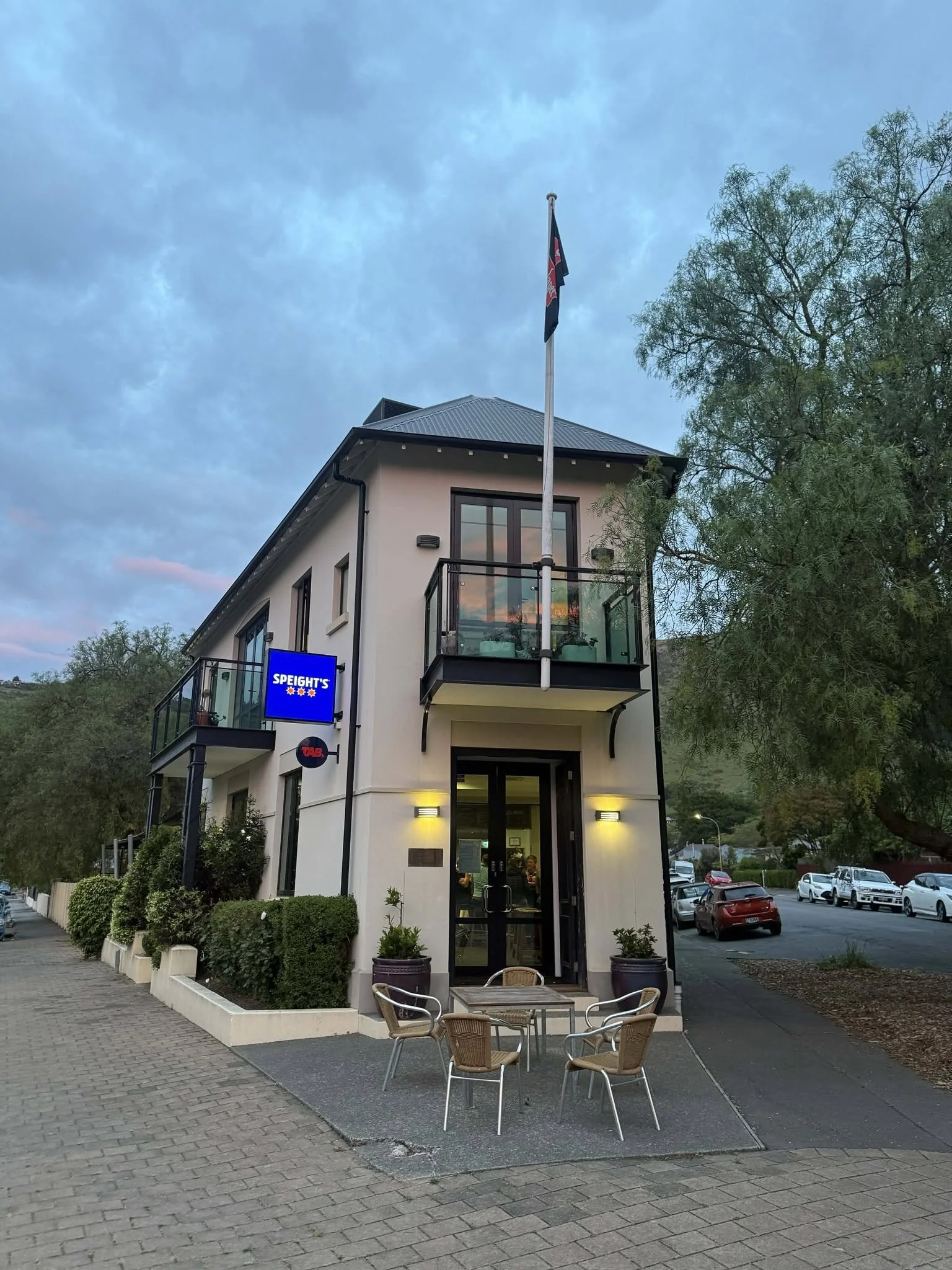 Speight's and TAB-branded pub building with outdoor patio seating, flagpole, trees, and parked cars at dusk.