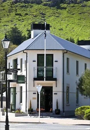 White two-story building with a gray metal roof beside a green hillside and street signs