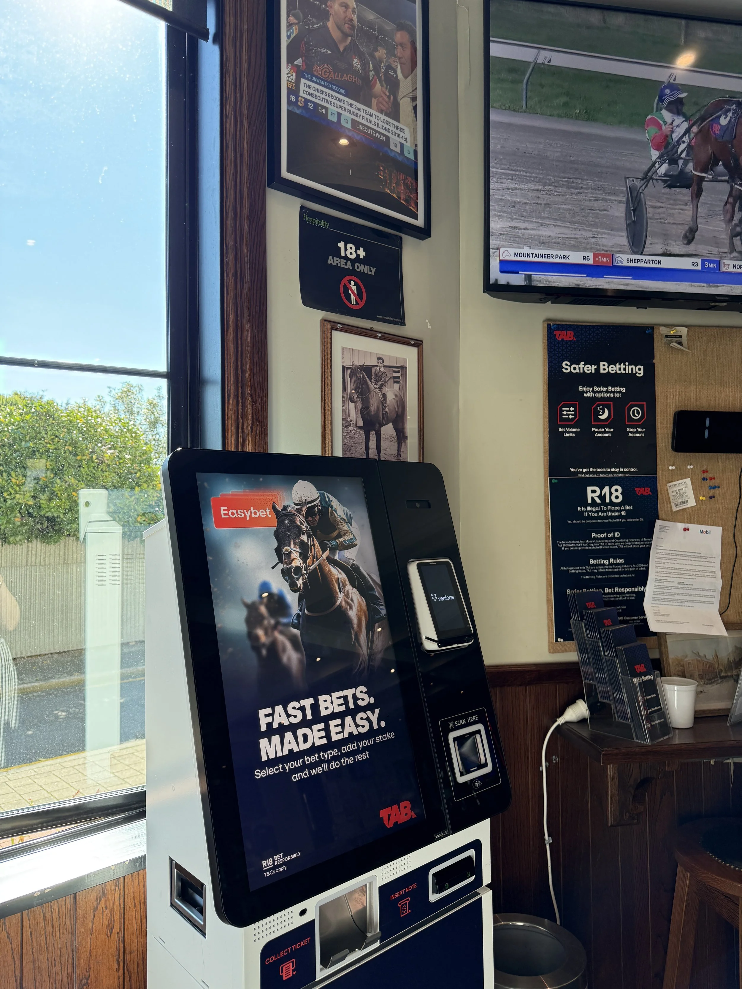 Interior of a TAB betting venue with a horse racing screen, betting kiosk, racing posters, and responsible gambling signs.