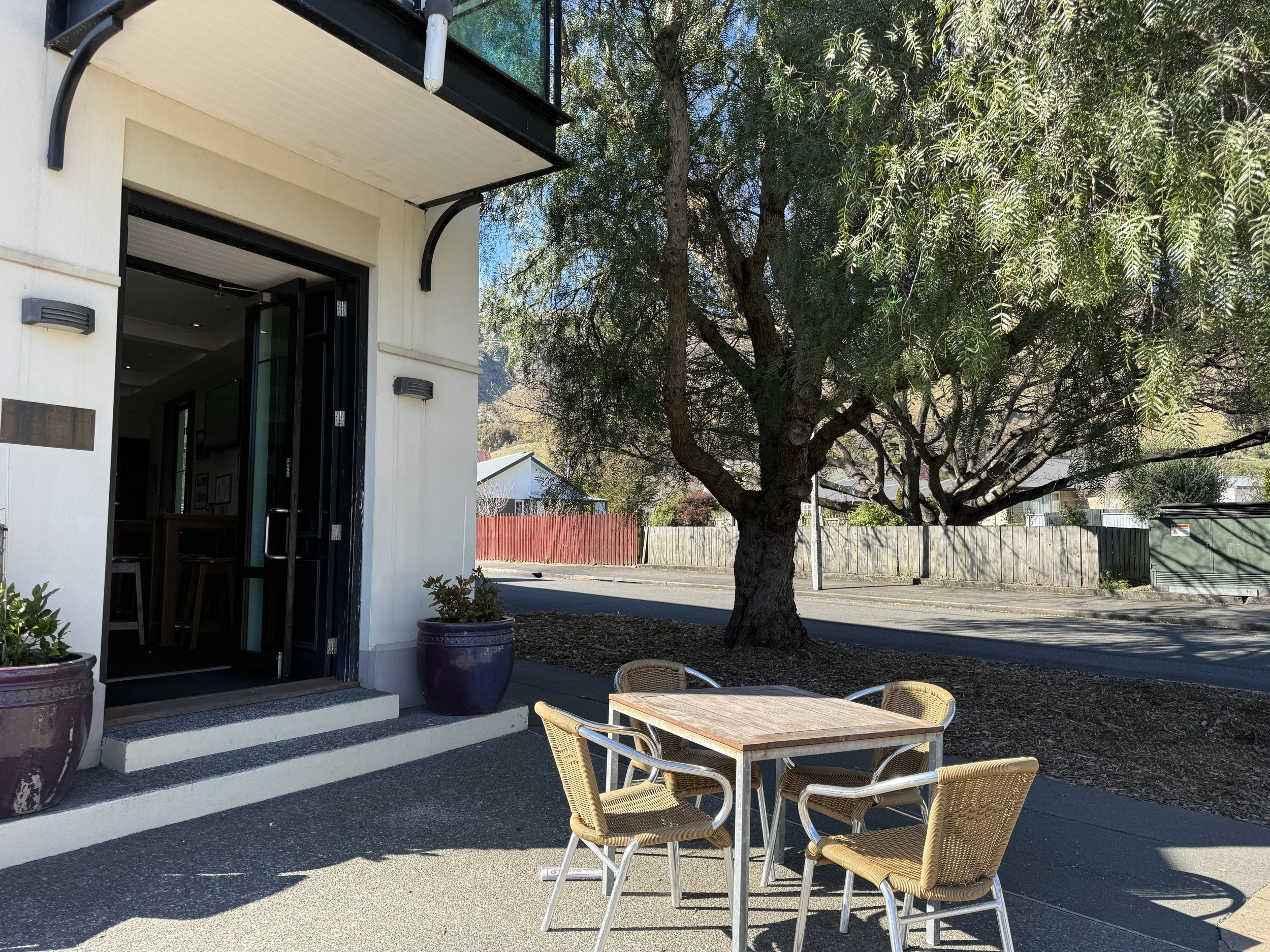 Outdoor patio with wicker chairs and a wooden table beside a building entrance, shaded by a large leafy tree.
