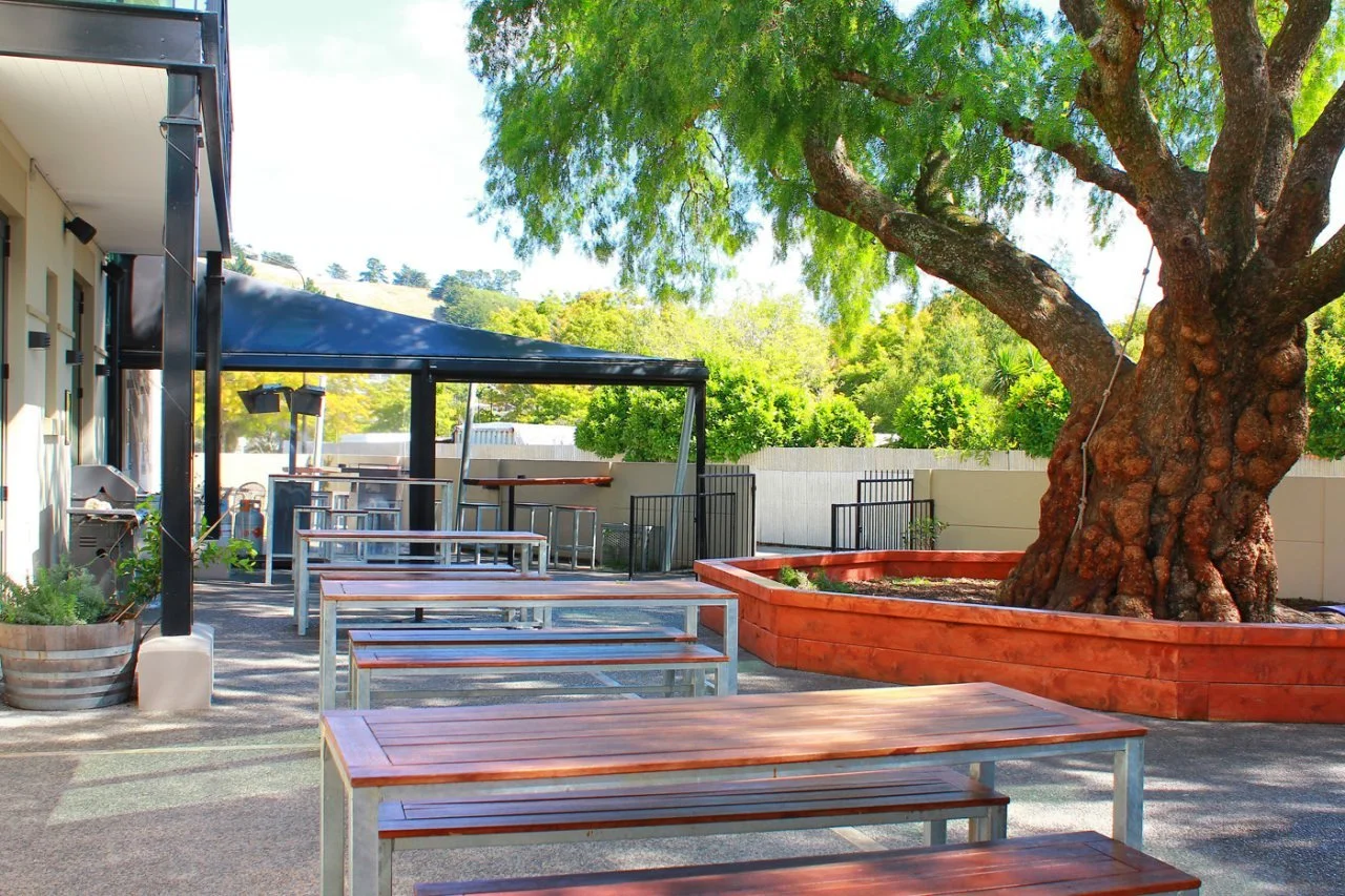 Outdoor patio with wooden picnic tables, a covered seating area, and a large tree in a raised planter
