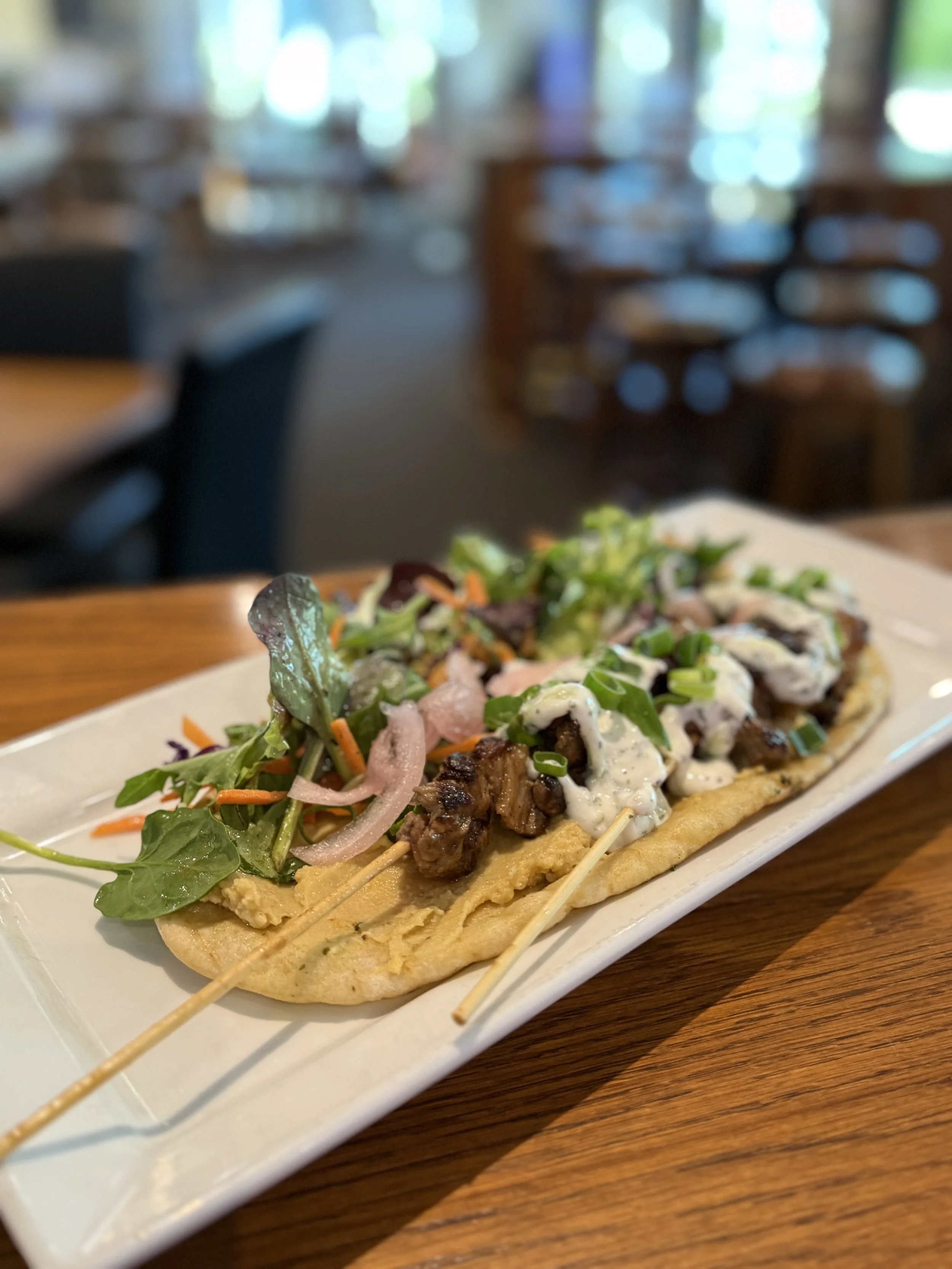 Grilled beef skewers with creamy sauce, flatbread, and mixed greens salad on a white plate