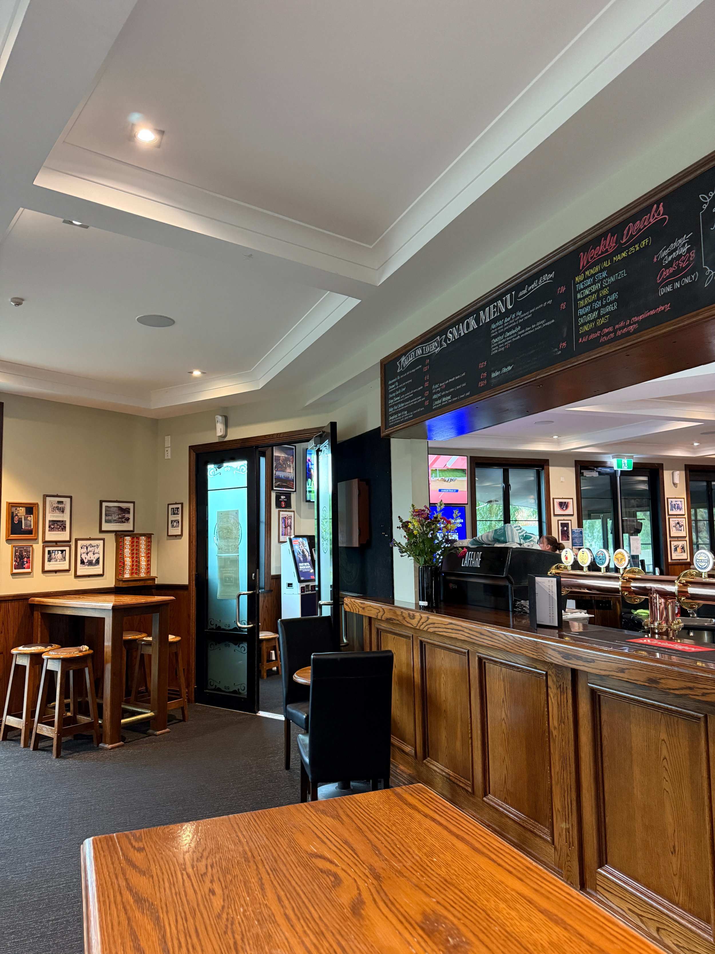 Interior of a traditional pub with a wooden bar, beer taps, chalkboard menus, framed wall photos, and high tables.
