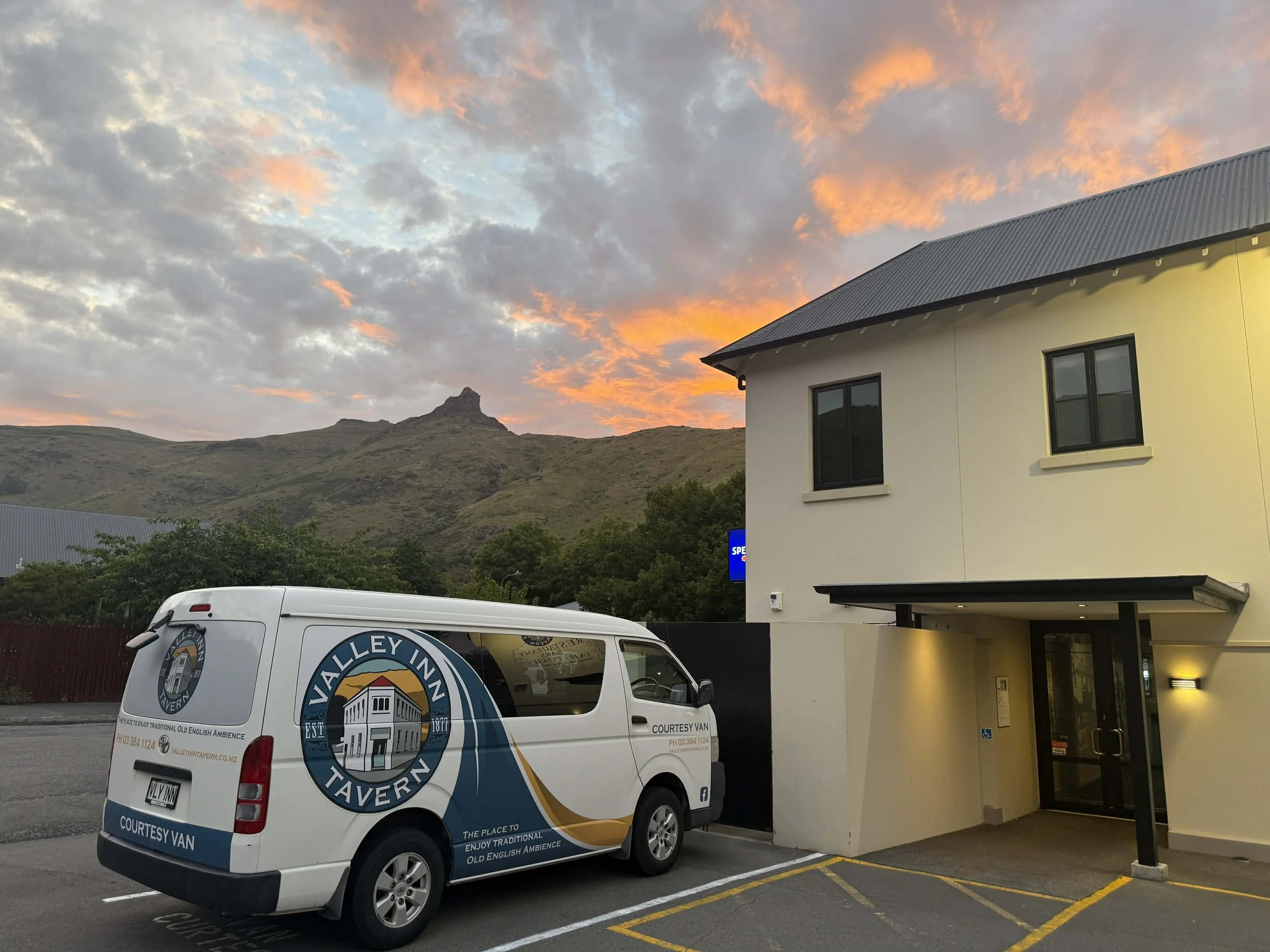 Valley Inn Tavern courtesy van parked outside a building with a rugged mountain backdrop under a colorful sunset sky