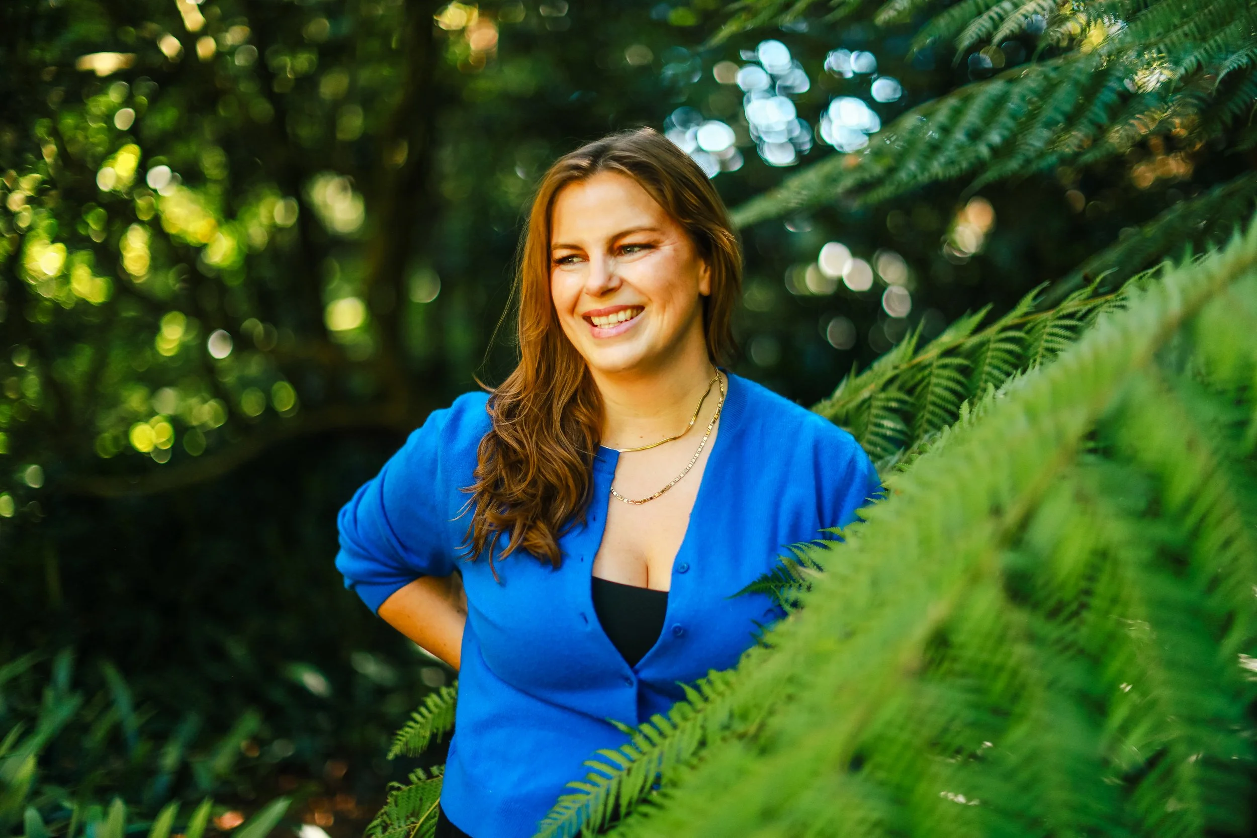 Lindsey is standing among lush green ferns and foliage.  She is smiling warmly and wearing a bright blue cardigan .