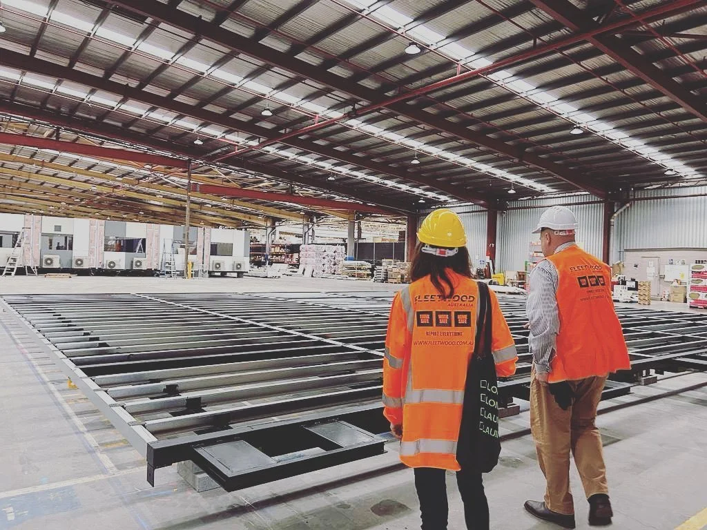 Two construction workers in safety vests and hard hats inspecting metal framework in a large warehouse under construction.