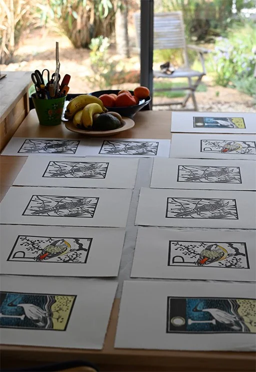 Table displaying Katy Hood's printed artworks, fruit, and a cup of drawing tools beside a garden window