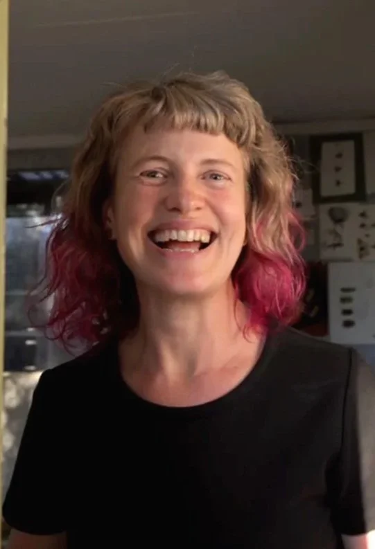 Katy Hood smiling woman with short brown and pink hair wearing a black dress in her studio entrance