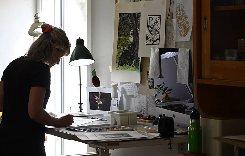 Katy Hood Artist working in her studio at a desk surrounded by artwork, prints, tools, and a desk lamp