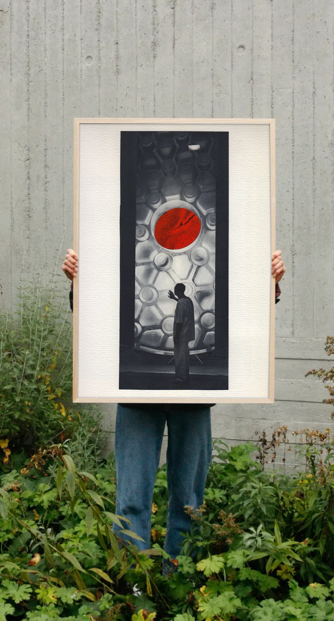 A person in a garden holds up a large framed print of a paper collage by Amanda Clifford, which features the silhouette of a man in scrubs holding out his hand to a futuristic structure. There is a window above which reveals a bright red planet