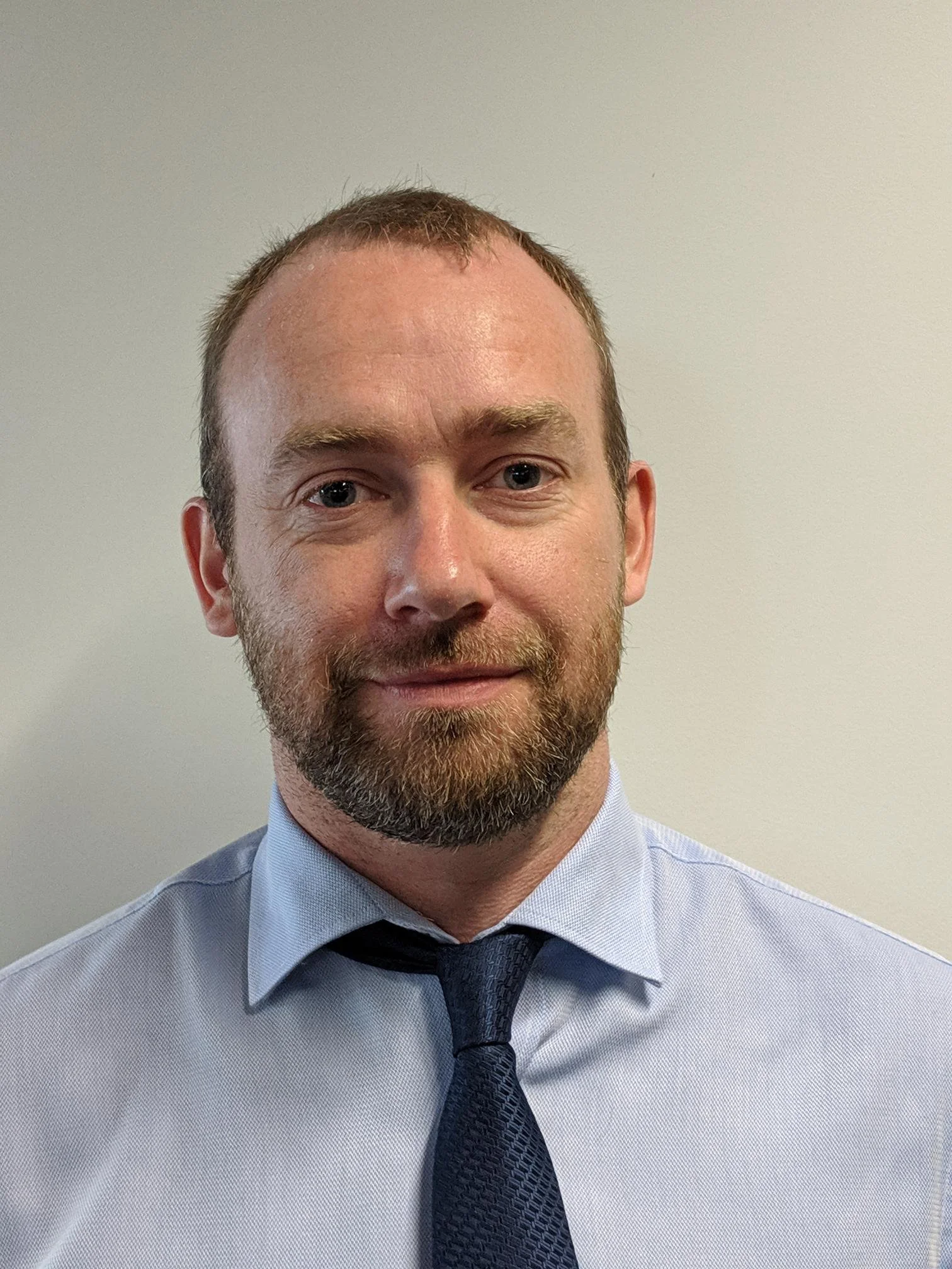A close-up photograph of a man wearing a light blue dress shirt, dark blue tie, and with a short beard and short brown hair, standing against a plain white wall.