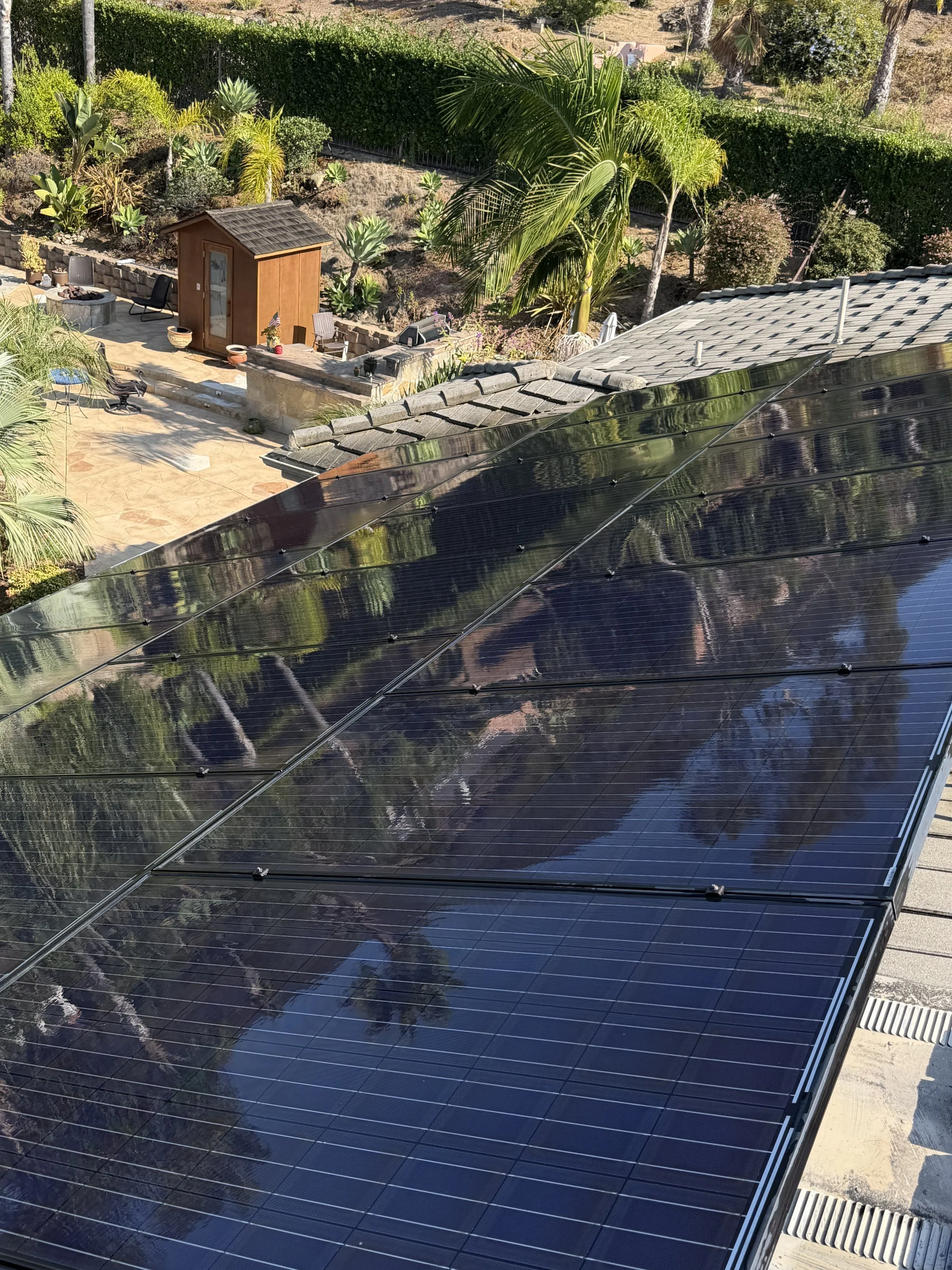 A backyard with solar panels installed on a roof in the foreground, and a patio area with outdoor furniture, a small shed, plants, trees, and landscape in the background.