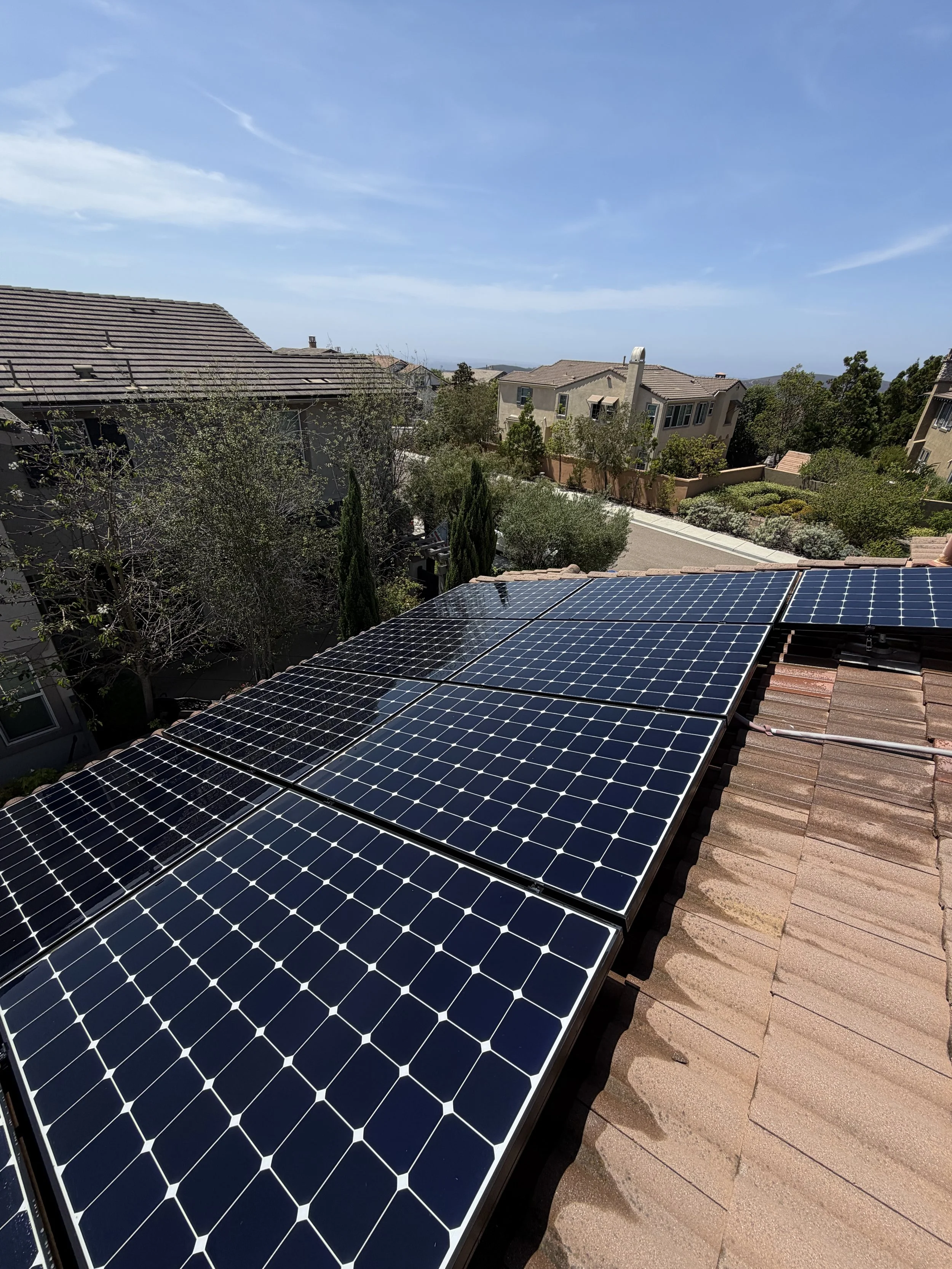 Solar panels installed on a residential rooftop with houses and trees in the background under a partly cloudy sky.