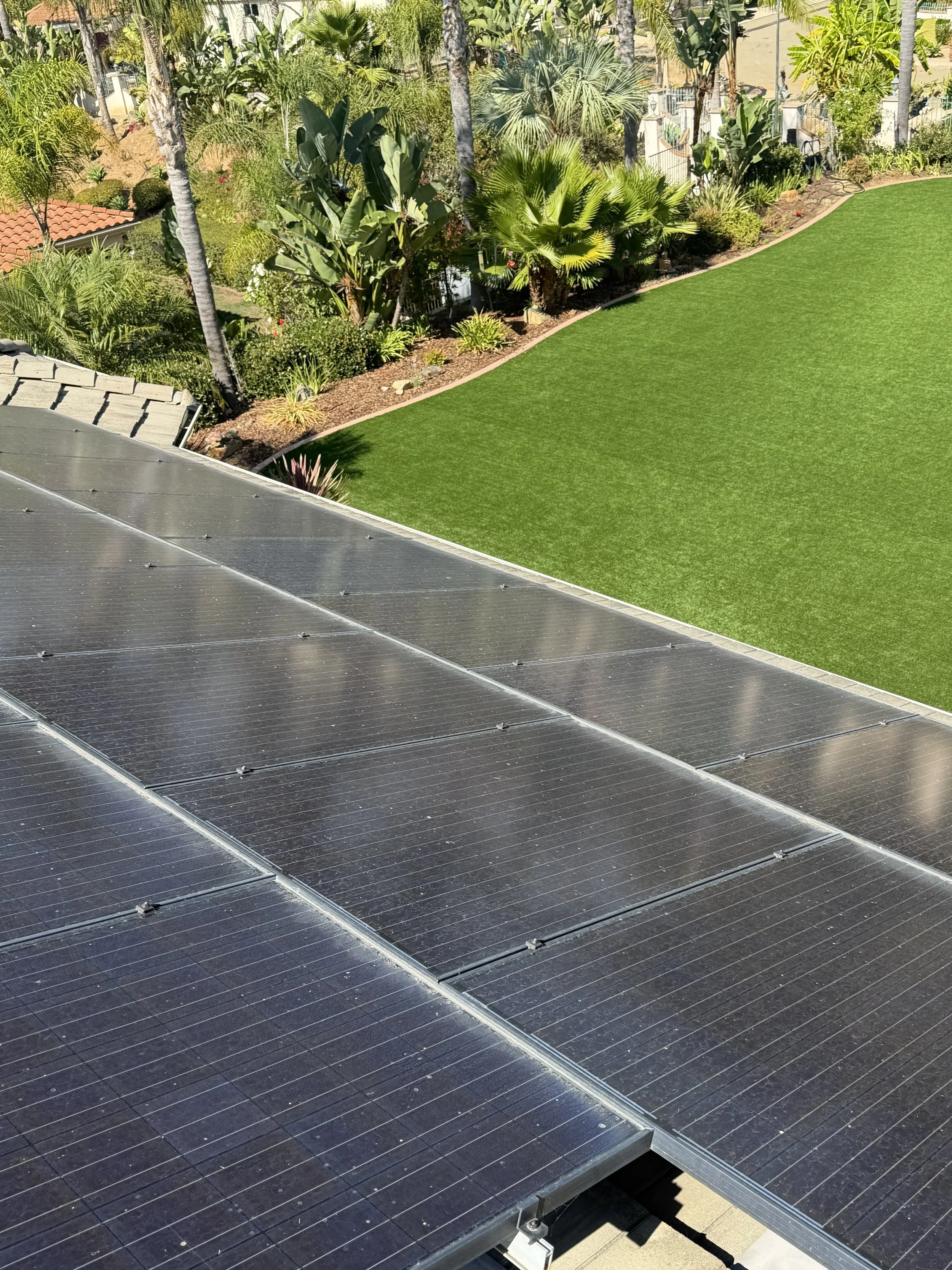 Solar panels installed on a roof above a green landscaped yard with trees and plants.