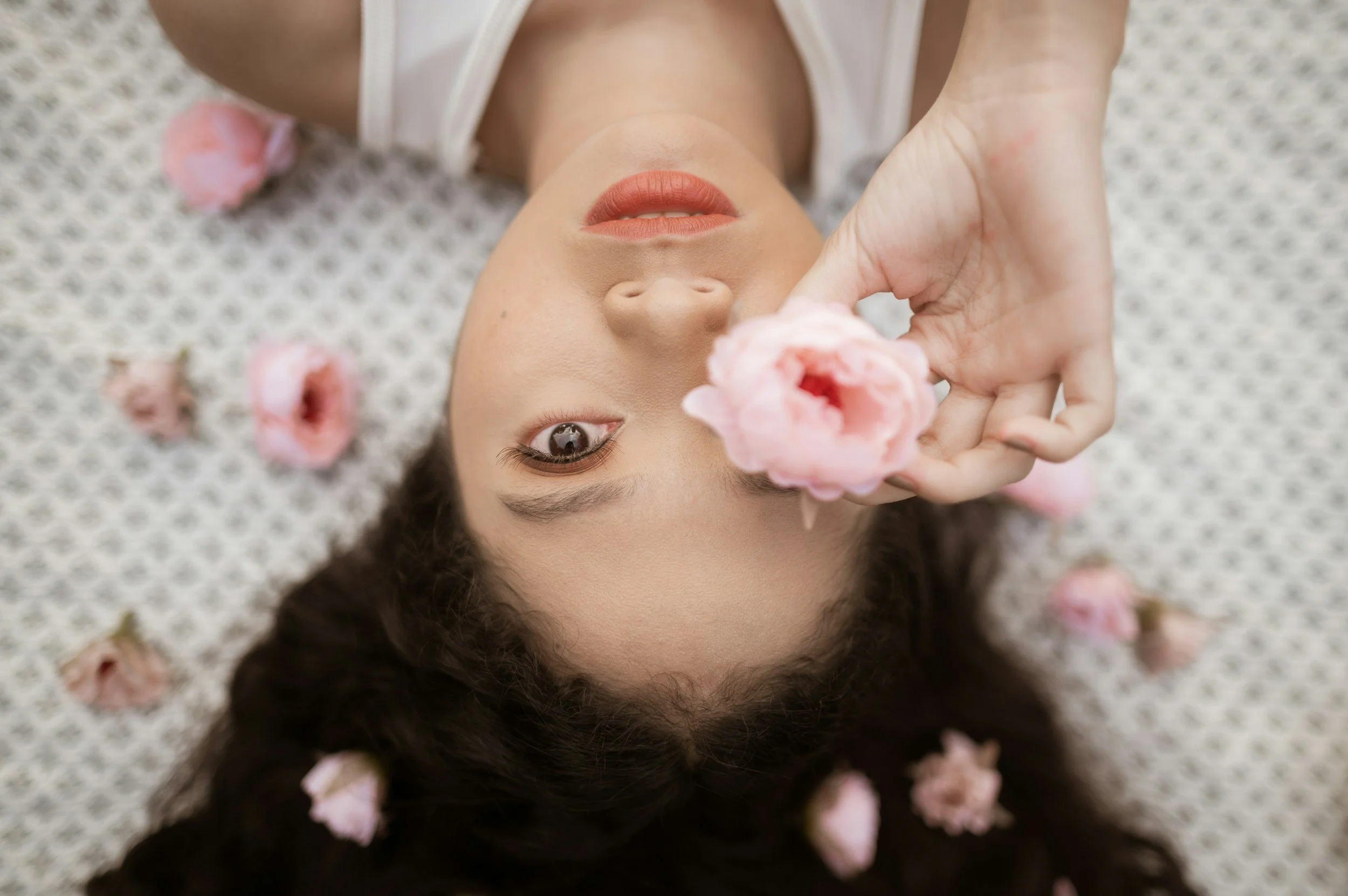 A woman lying on a textured surface surrounded by pink flowers, holding a flower near her eye, with her face partially visible and hair spread out.