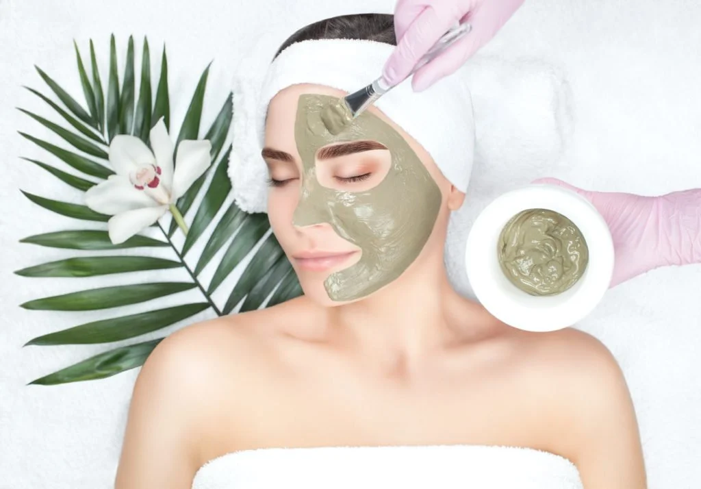 A woman receiving a facial treatment with a scrub or mask applied to her face, lying down with a towel wrapped around her head, next to a flower and green leaves.