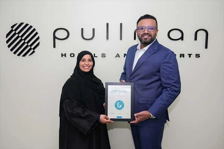 A man and woman, both smiling, holding a framed certificate in front of a wall with the logo 'Pullman Hotels and Resorts'.