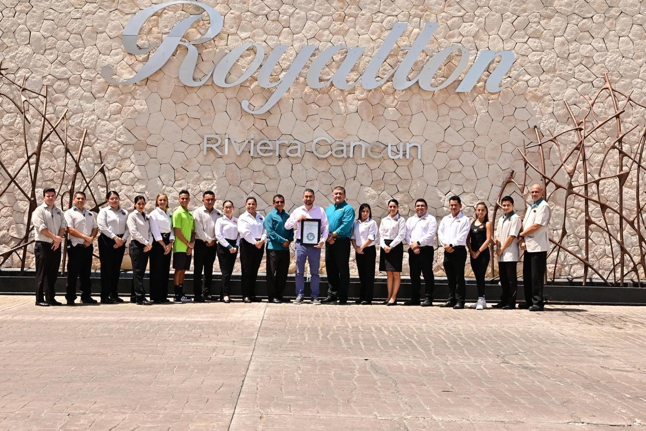 Group of people standing outside in front of a wall with the word "Riviera Cancun" on it. The group includes men and women dressed in business casual attire, some in white shirts and black pants, others in colorful shirts and black skirts or pants. One person in the middle is holding a framed certificate. The setting appears to be sunny and warm.