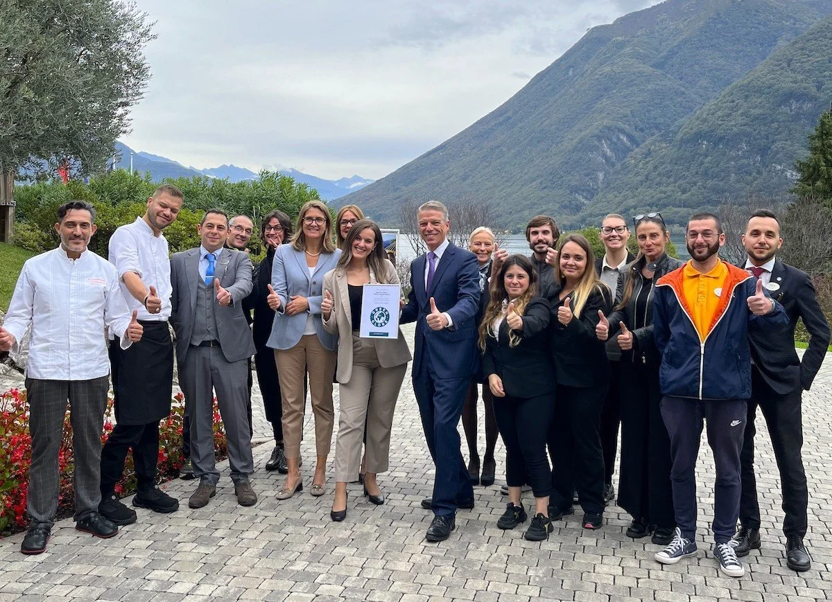 A group of diverse professionals standing outdoors on a cobblestone path with mountains and trees in the background. They are smiling and giving thumbs-up gestures.