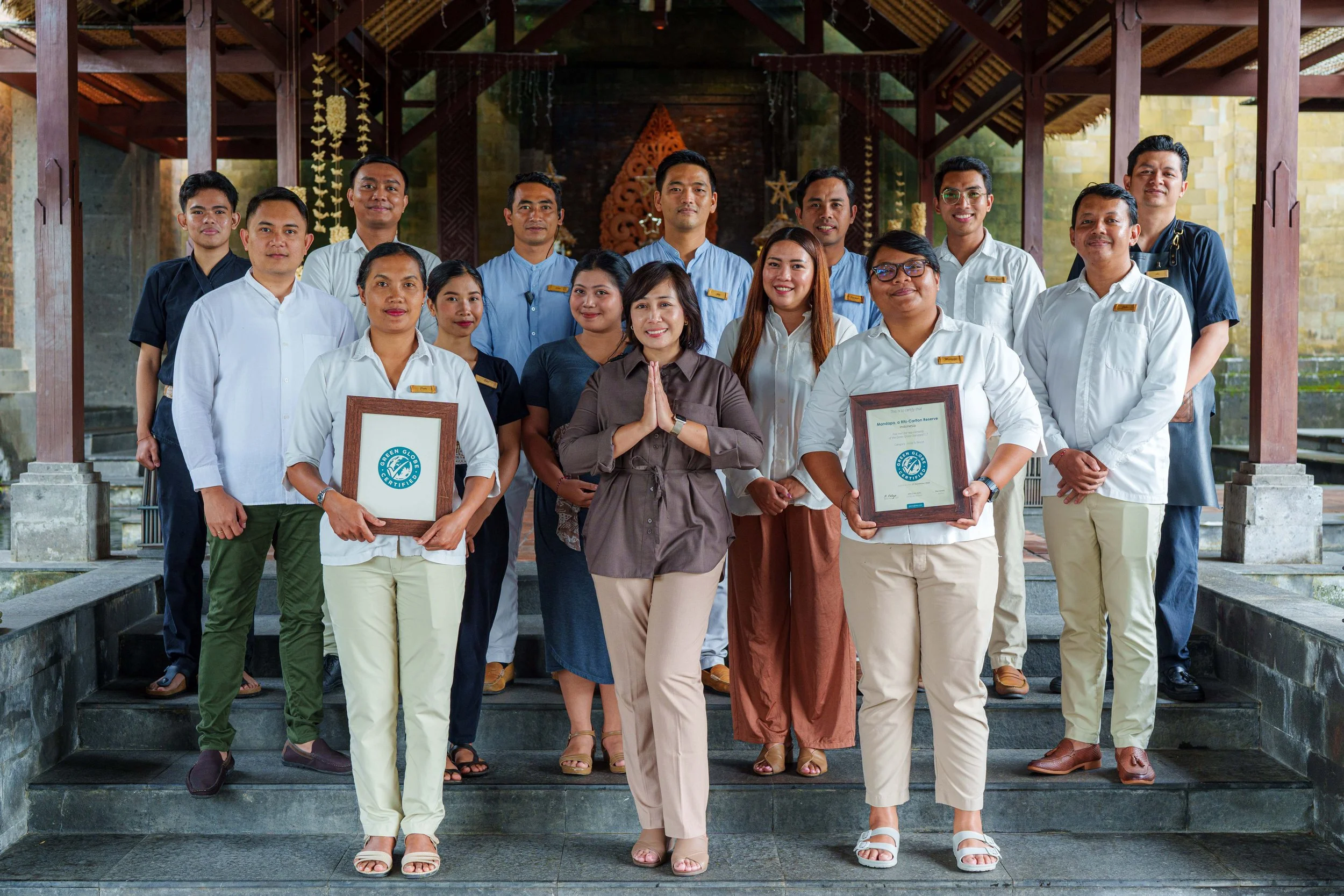 Group of 15 people standing on steps in front of wooden structure, some holding awards or certificates, posing for a photo.