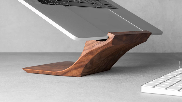 Laptop on a wooden stand on a gray desk with a white keyboard nearby.