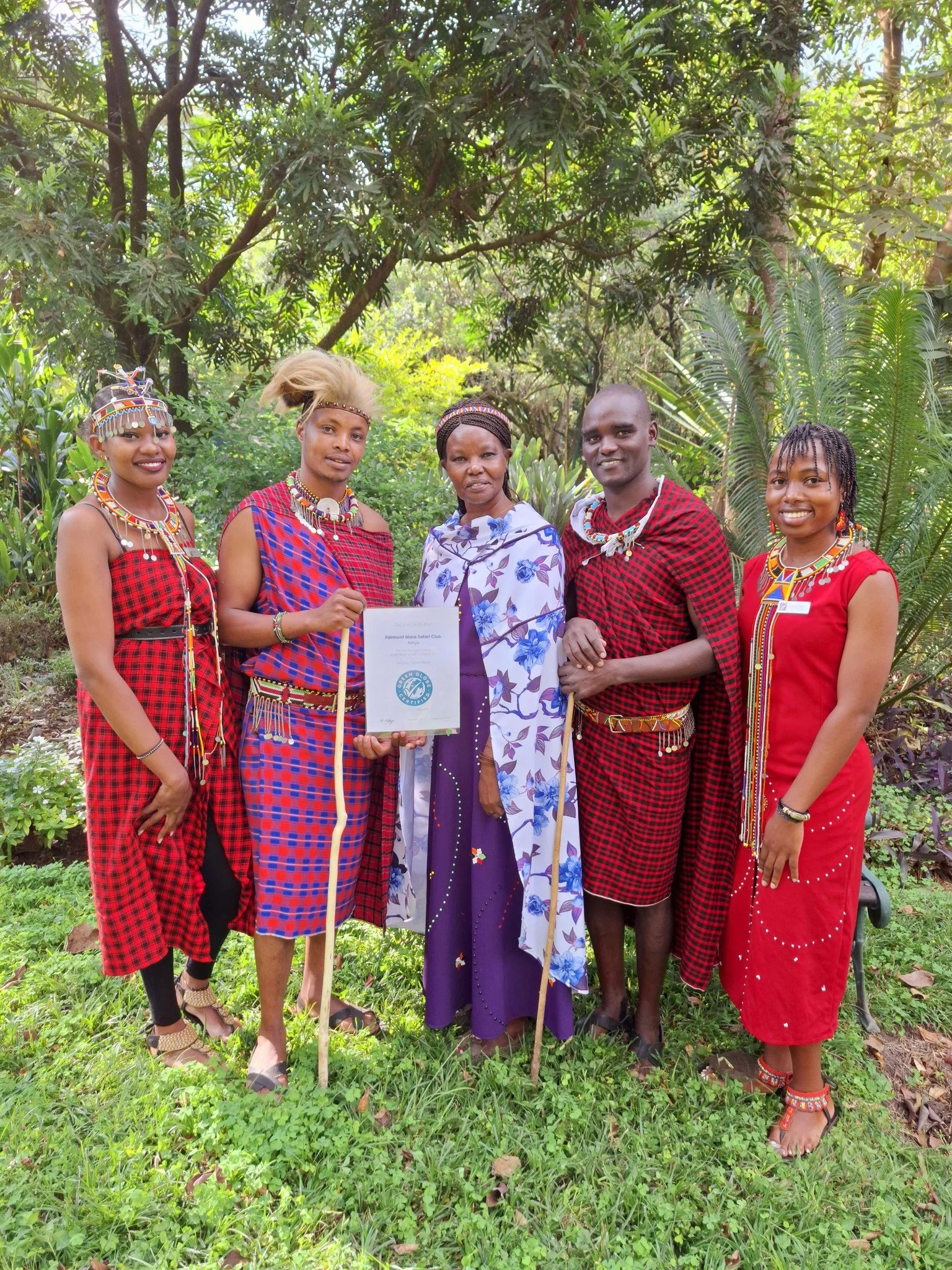 Group of five people in traditional Maasai attire standing outdoors with lush green trees in the background, holding a certificate.