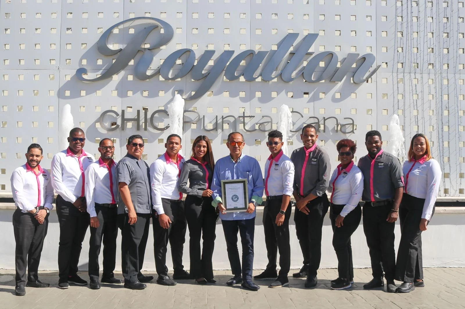 Group of fifteen people standing in front of a building with a sign that reads 'Fountain Chic Punta Cana'. They are dressed in business casual attire, some wearing uniforms with pink accents, and are smiling at the camera. One person in the center holds a framed certificate.