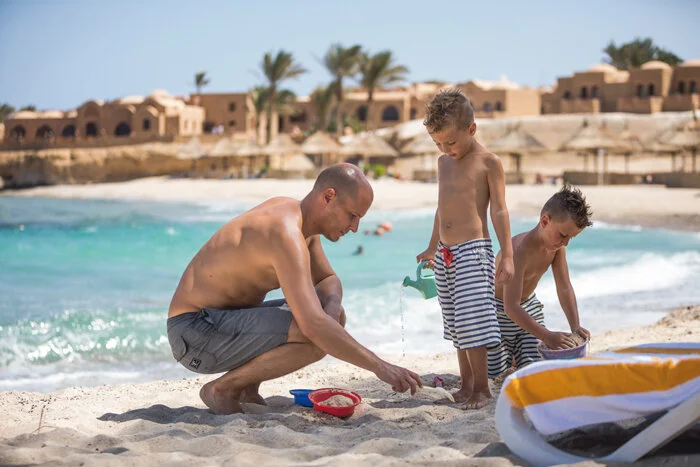 A man and two boys playing with sand and water on a beach, with ocean waves and buildings with palm trees in the background.