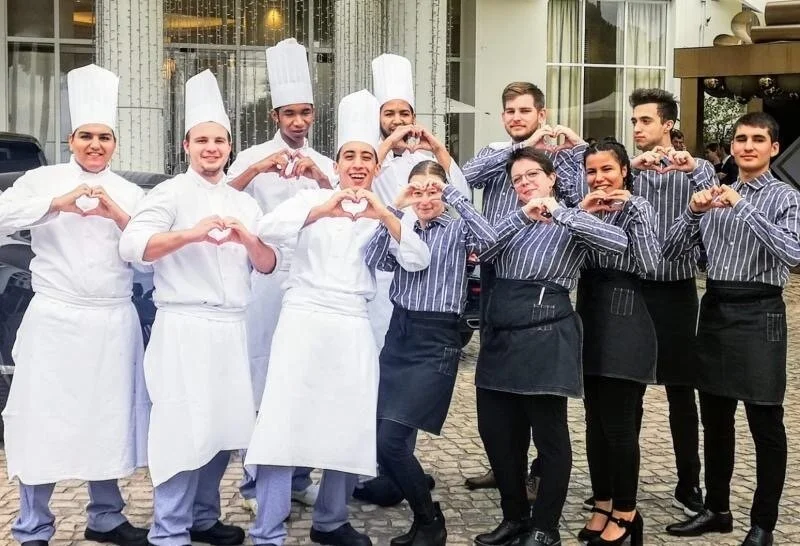 Group of chefs and waitstaff outside, all making heart shapes with their hands, dressed in white chef uniforms and striped shirts with black aprons.