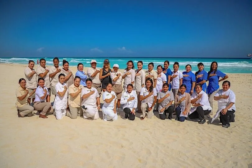 Group of people standing and kneeling on a sandy beach, with the ocean and a clear blue sky in the background.