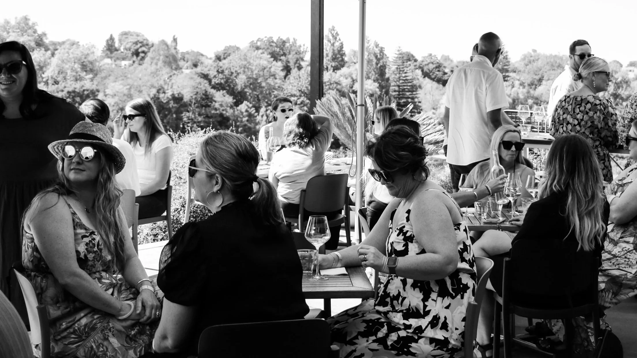 Group of people at an outdoor social gathering, some seated at tables with drinks, others standing, some wearing sunglasses and summer attire, with trees in the background.