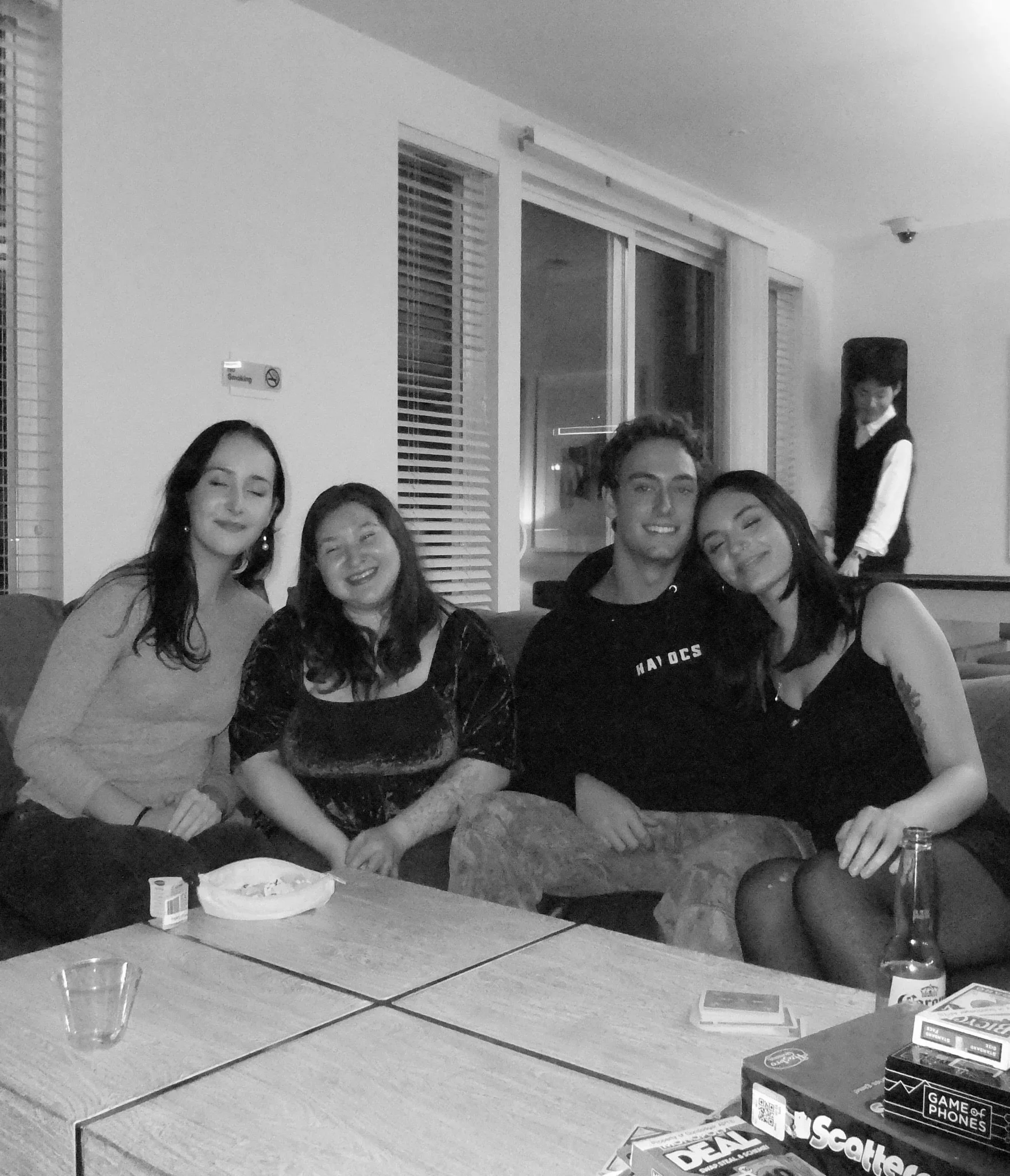 A group of five people sitting at a table inside a room, with snacks and drinks on the table, and a woman standing in the background near the wall.